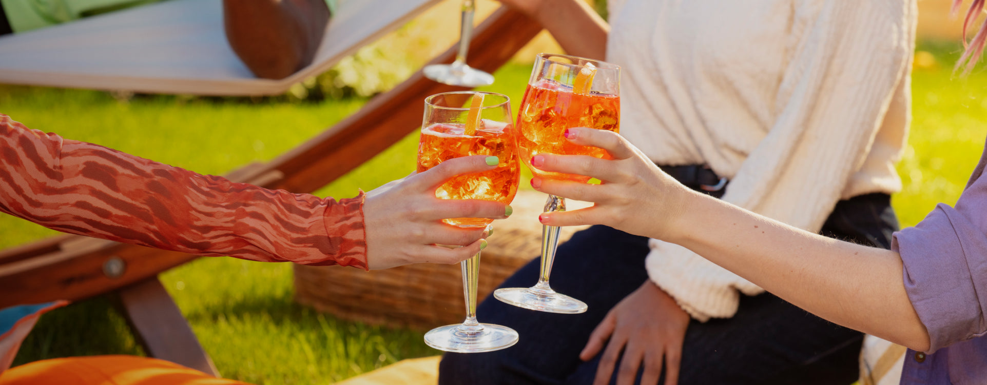 Three glasses of Aperol Spritz being clinked together by female hands in a sunny garden setting with a lounge chair and green grass in the background.